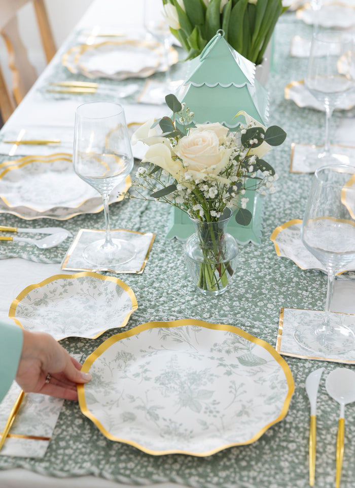A hand sets a Sophistiplate Timeless Sage Dinner Plate (8pk) with a gold rim on a green patterned tablecloth, surrounded by matching disposable plates, gold cutlery, wine glasses, floral napkins, and a vase of white flowers.