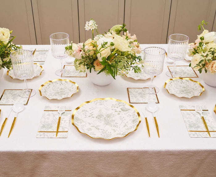 A beautifully set dining table features Sophistiplate’s Timeless Sage Dinner Plates, gold utensils, white napkins, clear glasses, and a centerpiece of white and pale yellow flowers in a white vase.