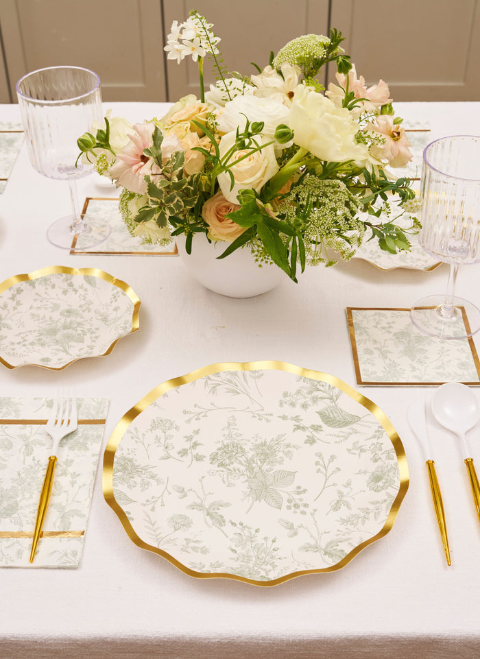 Elegant table setting with Sophistiplate Timeless Sage Dinner Plates, gold-trimmed napkins, clear glasses, gold-colored cutlery, and a centerpiece of white and blush flowers in a white vase on a white tablecloth.
