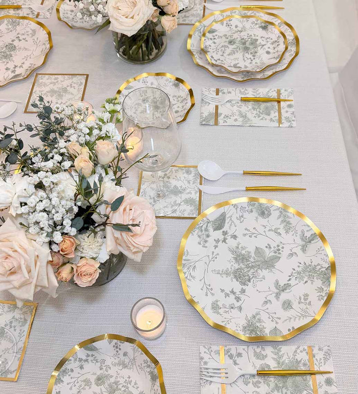 Table set for a meal with Sophistiplate Timeless Sage Dinner Plates, gold-edged cutlery, napkins, floral centerpieces of pale pink roses and greenery, glassware, a lit candle, all on a light-colored tablecloth.