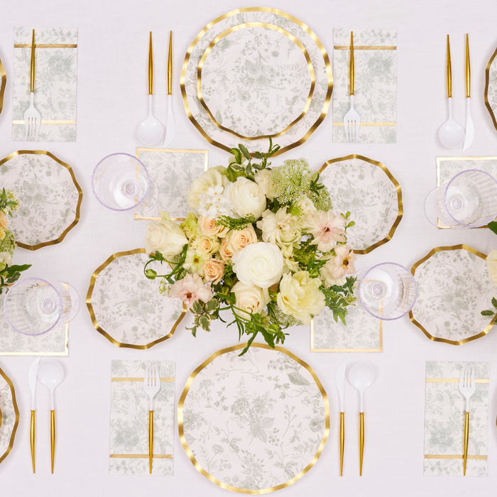 Elegant table setting with gold utensils, floral napkins, and cream-yellow flowers on a white tablecloth. Timeless Sage Salad Plates by Sophistiplate and disposable dinnerware add charm to the symmetrical decor.