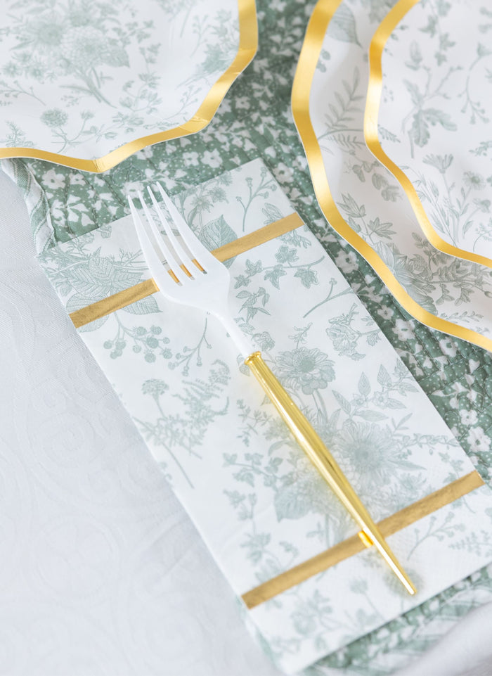 The Sophistiplate Timeless Table Setting: Sage features a white fork with a gold handle on a floral napkin with gold trim, next to designer disposable plates over a green and white tablecloth.