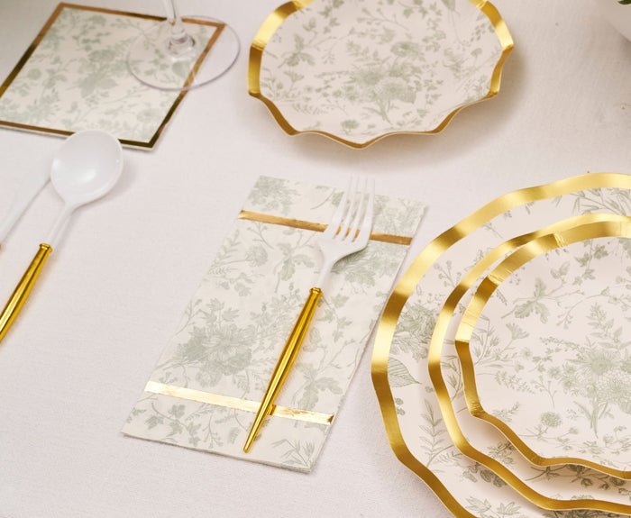 Elegant table setting with Sophistiplate Timeless Sage Guest Towels, gold-rimmed floral plates, gold and white utensils, and a wine glass on a gold-bordered coaster atop a white tablecloth.