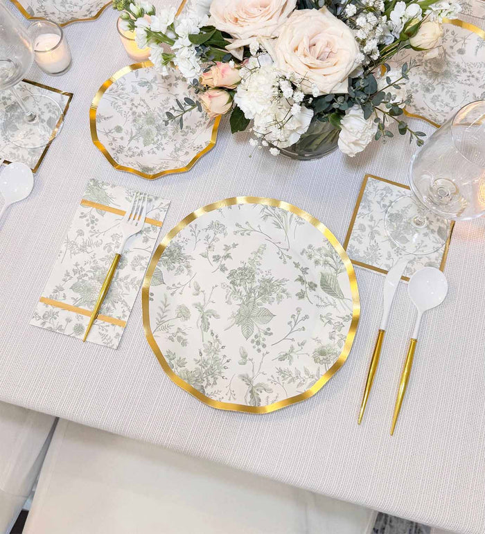 Elegant table setting featuring Sophistiplate Timeless Sage Dinner Plates (8 pack), gold-trimmed floral napkins, white plastic cutlery with gold handles, wine glasses, and a centerpiece of white and pale pink flowers.