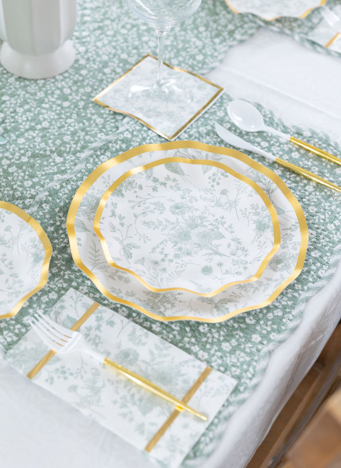 A close-up of the Timeless Table Setting: Sage by Sophistiplate shows designer disposable plates with floral patterns and gold trim, matching gold utensils, a green and white runner, and a wine glass on a coaster.