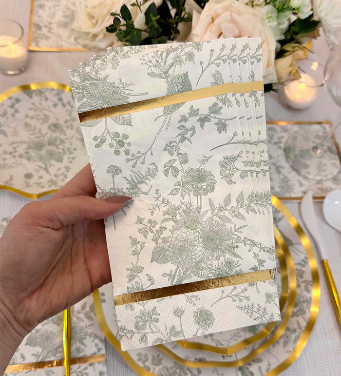 A hand holds Sophistiplate Timeless Sage Guest Towels (20pk) with gold foil trim above a table set with matching plates, gold utensils, white flowers, and candles.
