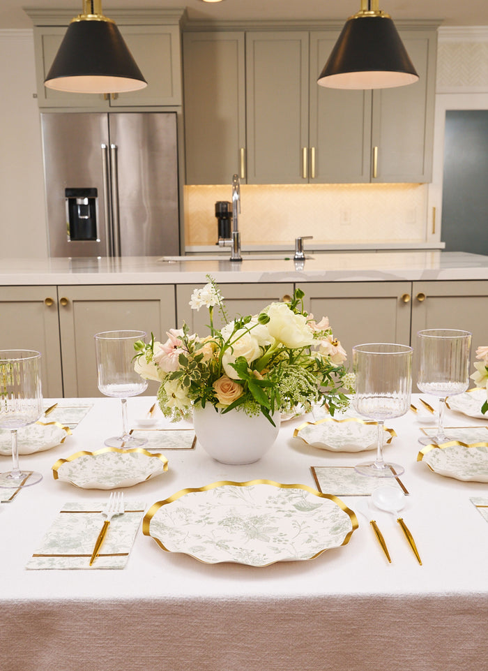 A modern kitchen table set features Sophistiplate Timeless Sage Dinner Plates (8pk), crystal glasses, floral napkins, and a white vase with white and pale pink flowers as the centerpiece.