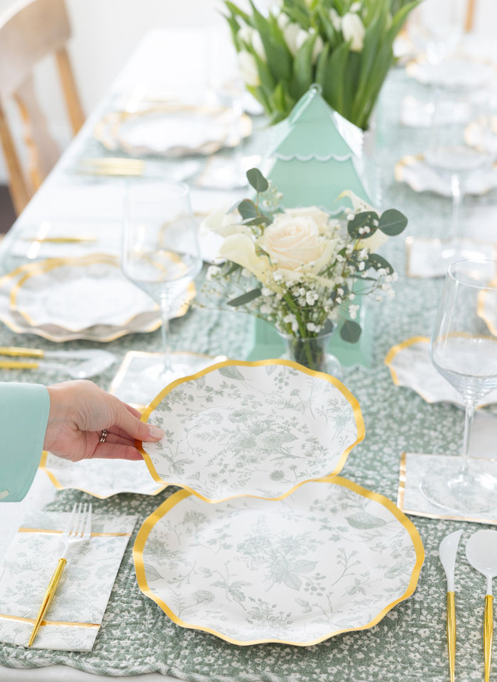 A hand holds a Sophistiplate Timeless Sage Salad Plate from an 8-pack, featuring a floral pattern and yellow rim, above a set dining table with matching plates, gold utensils, wine glasses, white roses, and a green tablecloth.