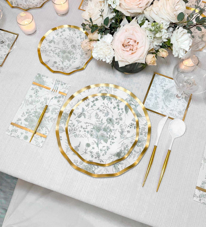 Elegant table setting with Sophistiplate Timeless Sage Salad Plates (8pk), matching napkins, gold flatware, and a centerpiece of white and blush flowers. Candles and clear glasses complete the look on a white tablecloth.