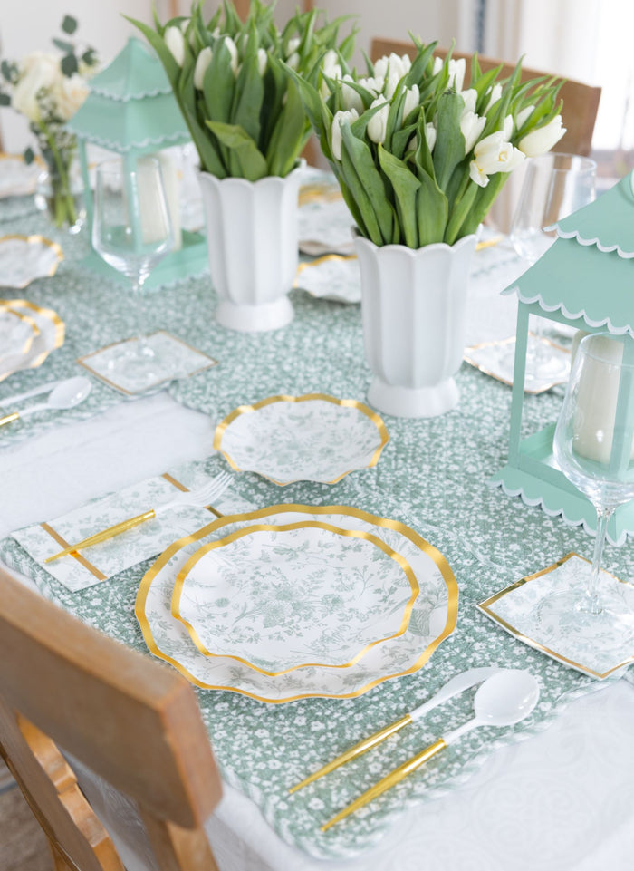 A spring-themed table features Sophistiplate Timeless Sage Salad Plates, gold flatware, and white napkins. White tulips in vases and pastel green lanterns sit atop a green and white tablecloth as charming centerpieces.