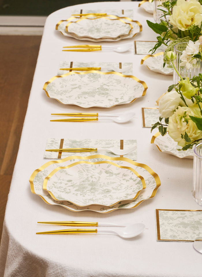 A neatly set table features Sophistiplate’s Timeless Table Setting: Sage—gold-rimmed designer disposable plates, gold-handled chopsticks, white plastic spoons, floral place cards, and a vase of white flowers.