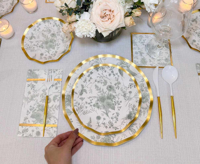 A hand arranges a Sophistiplate Timeless Sage Salad Plate from the 8-pack onto a set dining table with matching floral paper plates, gold utensils, centerpiece, wine glass, and candles—perfect for elegant disposable dinnerware.