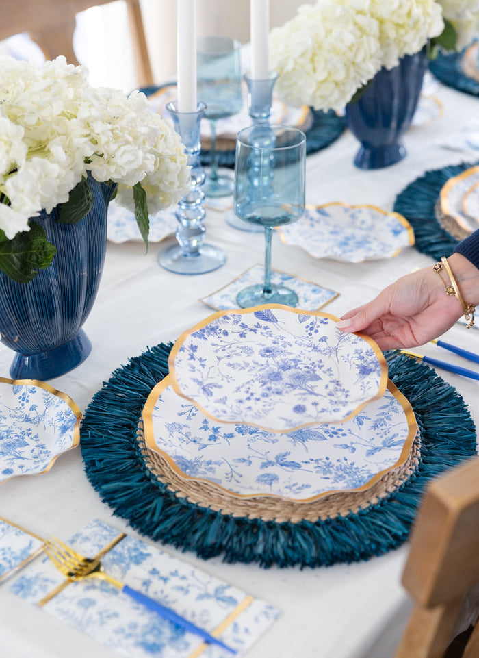 A hand sets a blue and white floral plate on a layered place setting with gold accents, blue glassware, and Sophistiplate Timeless Guest Towels, complemented by white flowers in blue vases on an elegant table.