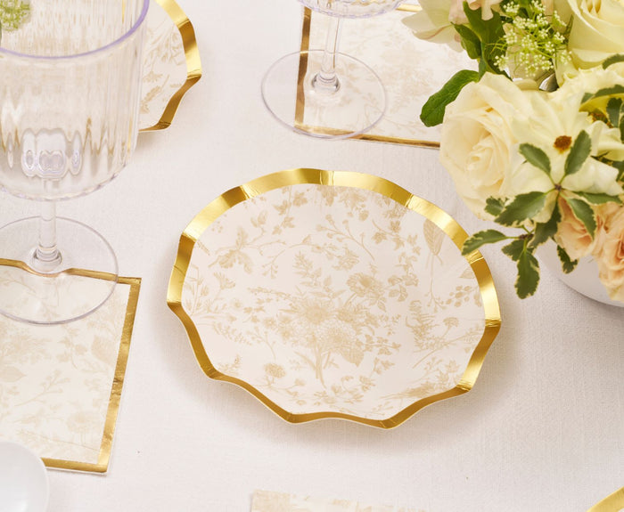 The Sophistiplate Timeless Sand Appetizer/Dessert Bowls (8pk) with scalloped gold edges and a floral design rest on a white tablecloth, surrounded by clear glassware, matching napkins, and a bouquet of cream roses.