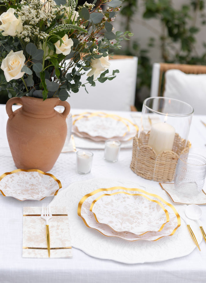 An outdoor table is set with white and gold plates, gold cutlery, white napkins, Sophistiplate Timeless Sand appetizer/dessert bowls, a terracotta vase of white roses, and a candle in a wicker holder on a crisp white tablecloth.