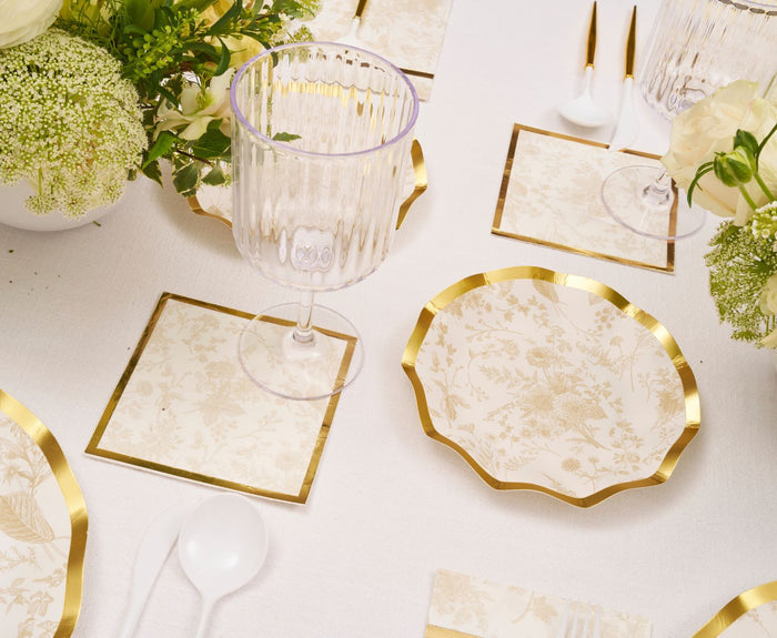 Elegant table setting with Sophistiplate Timeless Sand appetizer/dessert bowls (8pk), cream and gold floral plates, square coasters, clear ribbed wine glasses, white plastic cutlery, and floral centerpieces on a white tablecloth.