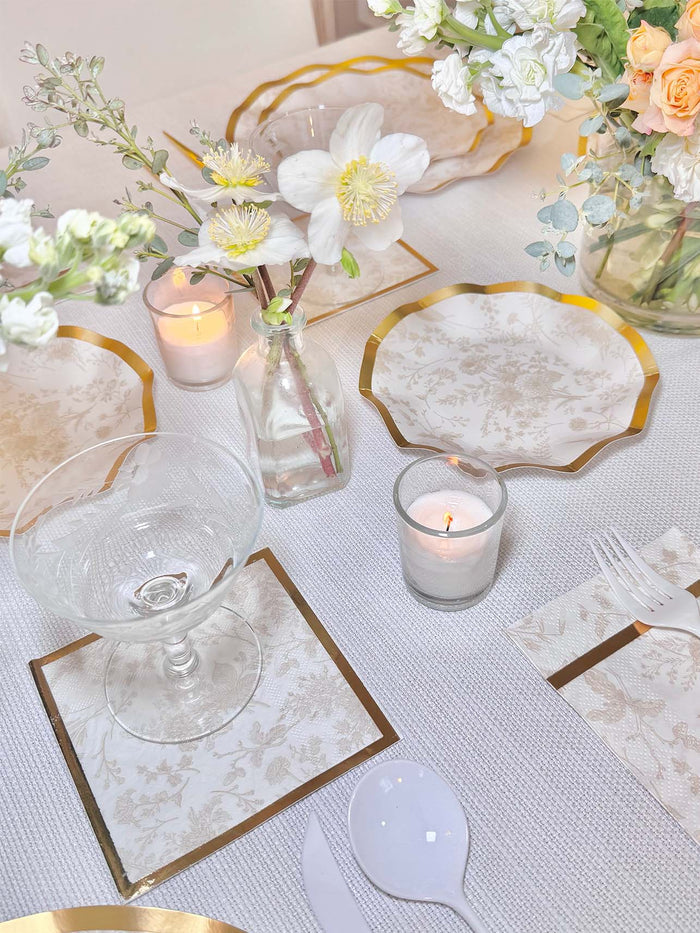 Elegant table setting with Sophistiplate Timeless Sand Appetizer/Dessert Bowls (8pk), gold-trimmed floral plates, glassware, white cutlery, vases of flowers, candles, matching napkins, and a white textured tablecloth.