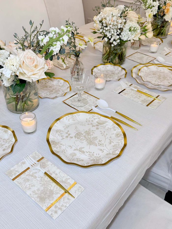 A beautifully set table showcases Sophistiplate’s Timeless Sand Dinner Plates (8pk), gold utensils, crystal glasses, white napkins, candles, and bouquets of white and blush flowers in glass vases on a crisp white tablecloth.