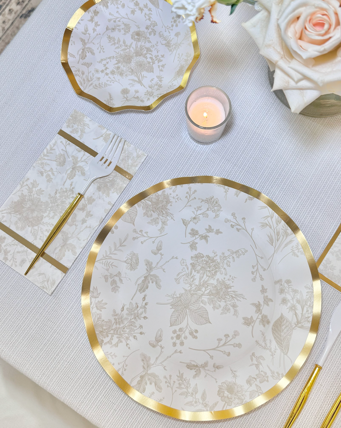 Create an elegant table setting with Sophistiplate’s Timeless Sand Dinner Plates (8pk), featuring gold floral scalloped edges, paired with gold flatware, a gold-accented napkin, candlelight, and a white rose centerpiece on a white tablecloth.