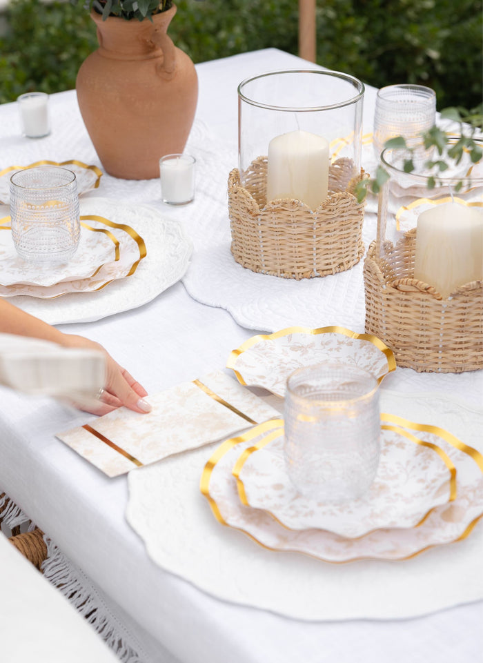 A hand places a Timeless Sand Guest Towel by Sophistiplate on a white outdoor table set with layered plates, clear glasses, wicker candle holders, and a terracotta vase for a serene and sophisticated tablescape.