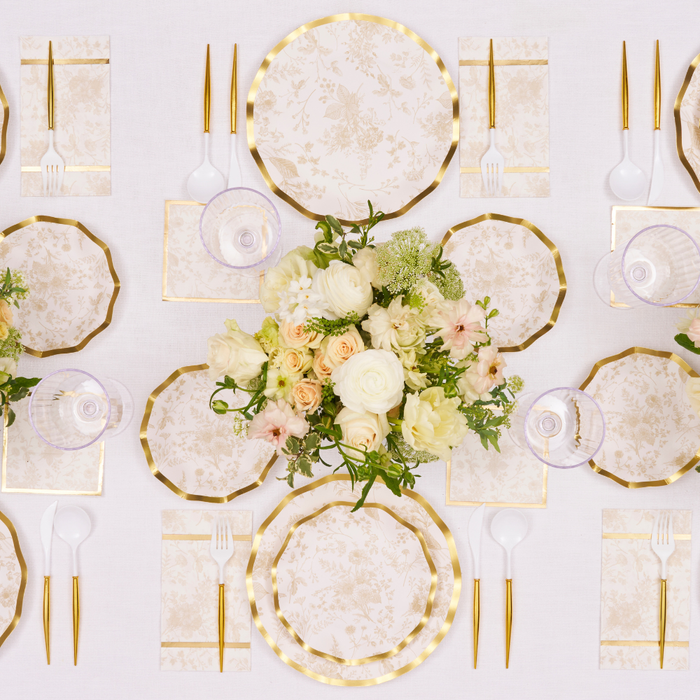 Sophistiplate's Timeless Sand Dinner Plates (8pk) create an elegant table set with gold flatware, crystal glasses, napkins, a white floral centerpiece, and a white tablecloth—arranged for a sophisticated celebration.