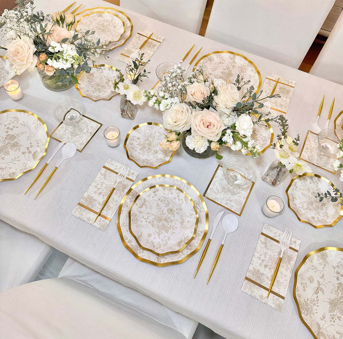 Elegant table setting with Sophistiplate's Timeless Sand Salad Plates (8pk), gold utensils, white napkins, floral centerpieces of white and pale pink blooms, and small candles—all atop a crisp white tablecloth.