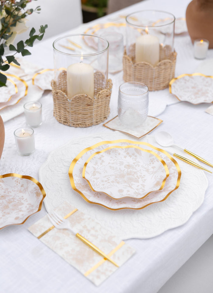 A close-up of the Timeless Table Setting: Sand by Sophistiplate, featuring designer disposable plates with gold trim, gold cutlery, clear glasses, woven candle holders, and white napkins on a white tablecloth.