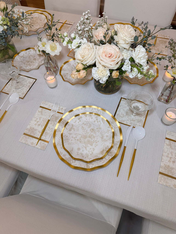 Elegant table setting with Sophistiplate Timeless Sand Salad Plates (8pk), gold utensils, white napkins, clear glasses, and a floral centerpiece of white and blush roses. Decorative candles and flower arrangements add a sophisticated touch.