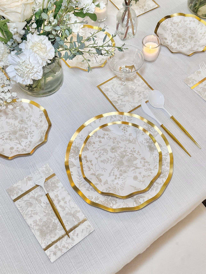 A formal table setting with Sophistiplate Timeless Sand Salad Plates (8pk), gold utensils, white flowers in glass vases, and lit candles arranged elegantly on a white tablecloth.