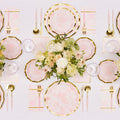 The Timeless Table Setting: Blush by Sophistiplate features pink and gold-rimmed heavy duty paper plates, gold cutlery, pink napkins, clear glasses, and a white and blush floral centerpiece on a white tablecloth.
