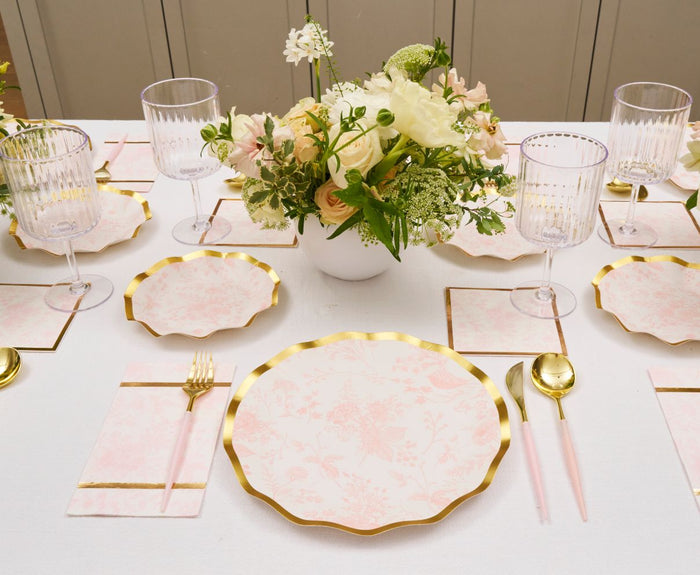 An elegant table set with Sophistiplate Timeless Blush Dinner Plates, gold cutlery, clear ribbed glasses, floral napkins, and a white floral centerpiece in a round vase creates a refined and light atmosphere.