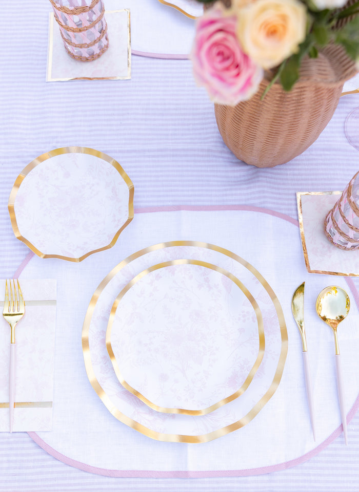 A pastel tablescape with gold cutlery, pink glasses, a white napkin, and a wicker vase of pink and yellow roses on a lilac-striped cloth, featuring Sophistiplate's Timeless Blush Salad Plates (8pk) for an elegant touch.