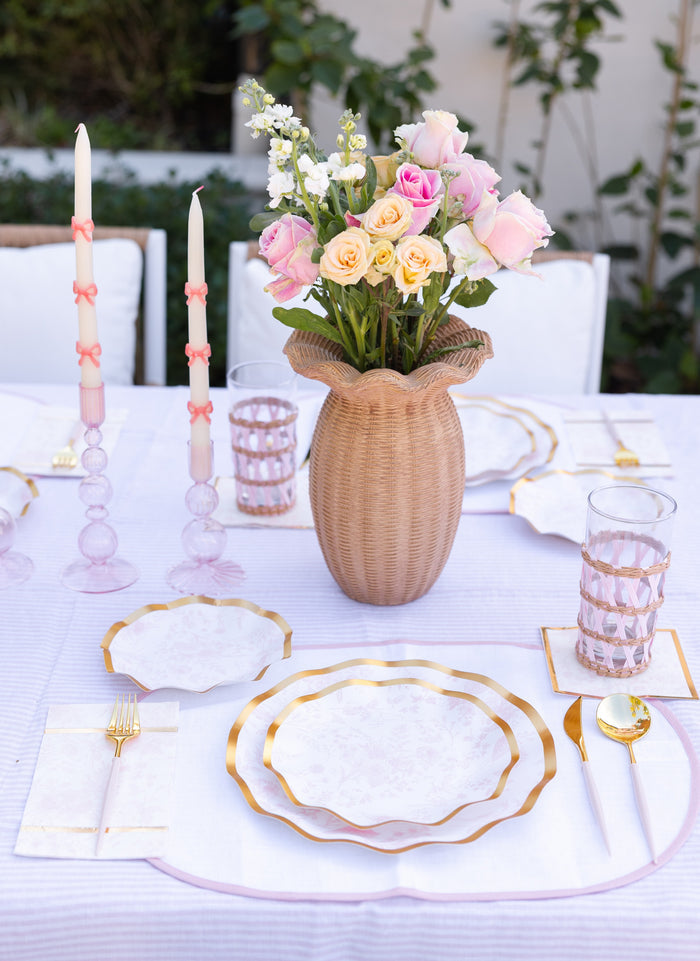 A table set for a meal with Sophistiplate Timeless Blush Dinner Plates, gold cutlery, pink glasses, pink candles in holders, and a woven vase with pink and yellow roses as the centerpiece. The tablecloth and napkins are white.