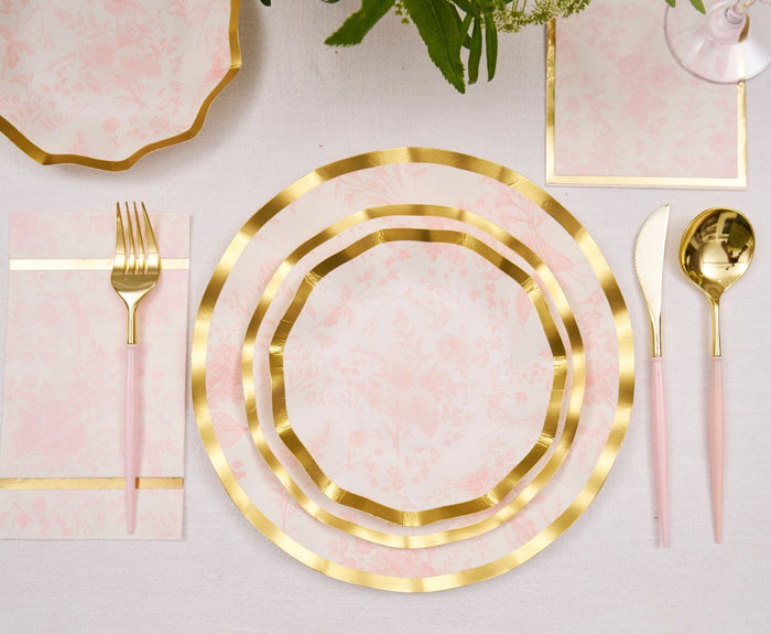 The Timeless Table Setting: Blush by Sophistiplate features gold-trimmed, pale pink floral heavy-duty paper plates, gold cutlery with pink handles, matching napkins, and a floral centerpiece—your perfect party-in-a-box.