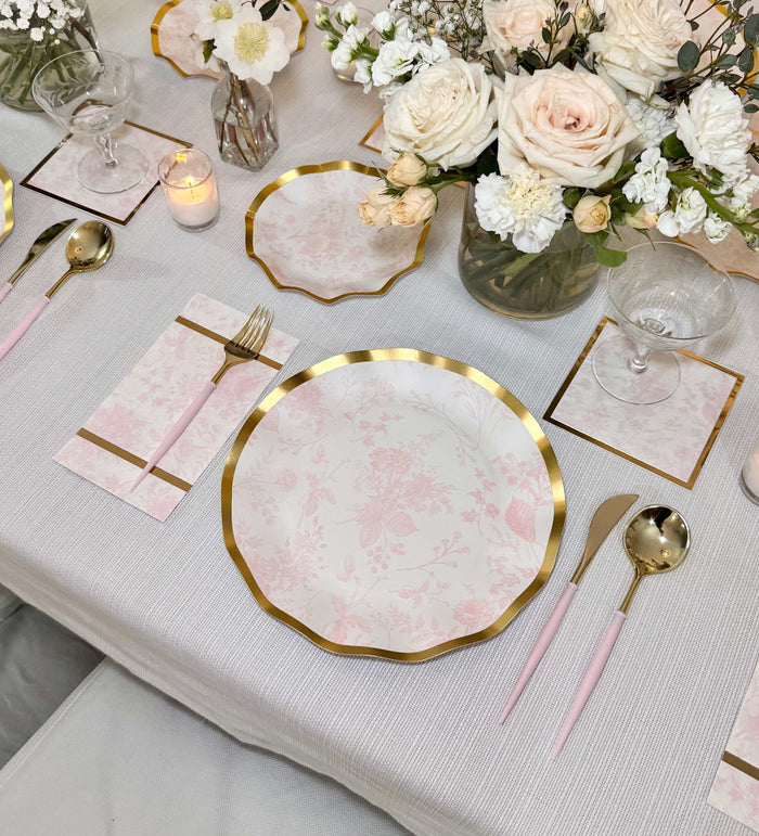 Create an elegant table setting with Sophistiplate Timeless Blush Dinner Plates (8pk), paired with pink and gold cutlery, floral napkins, crystal glasses, white florals, a lit candle, and small vases on a white tablecloth.