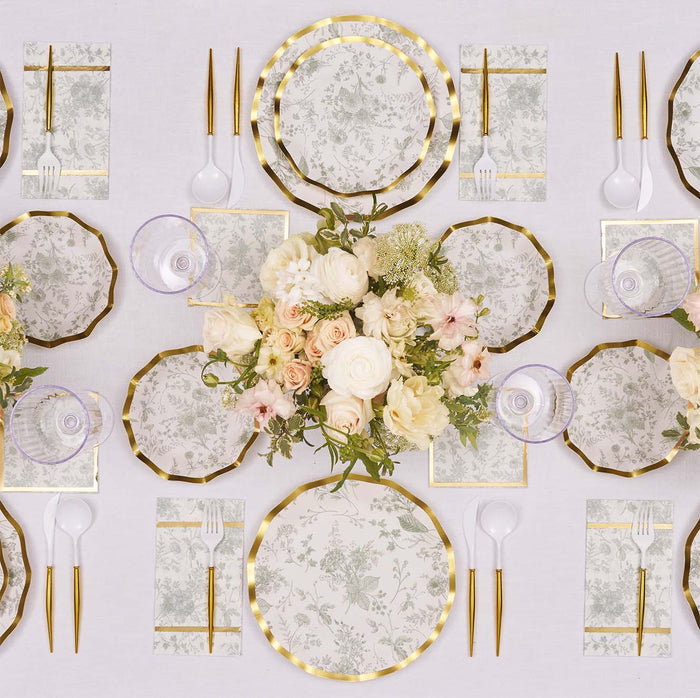 Experience an elegant table setting with Sophistiplate’s Timeless Table Setting: Sage, featuring designer disposable plates with floral patterns, gold-rimmed cutlery, napkins, clear cups, and a central arrangement of white and blush flowers on a white tablecloth—all arranged in a symmetrical display.