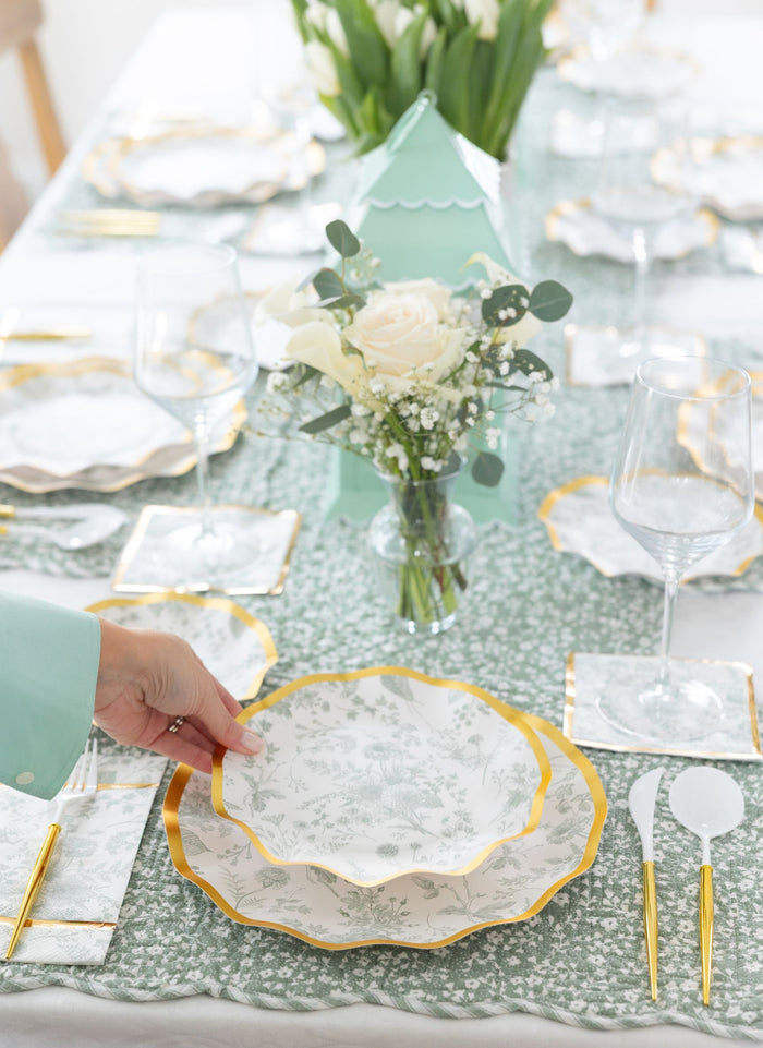 A hand sets a Sophistiplate Timeless Table Setting: Sage designer disposable plate with gold trim on a green and white tablecloth, surrounded by elegant plates, utensils, glasses, floral centerpieces, and a green lantern.