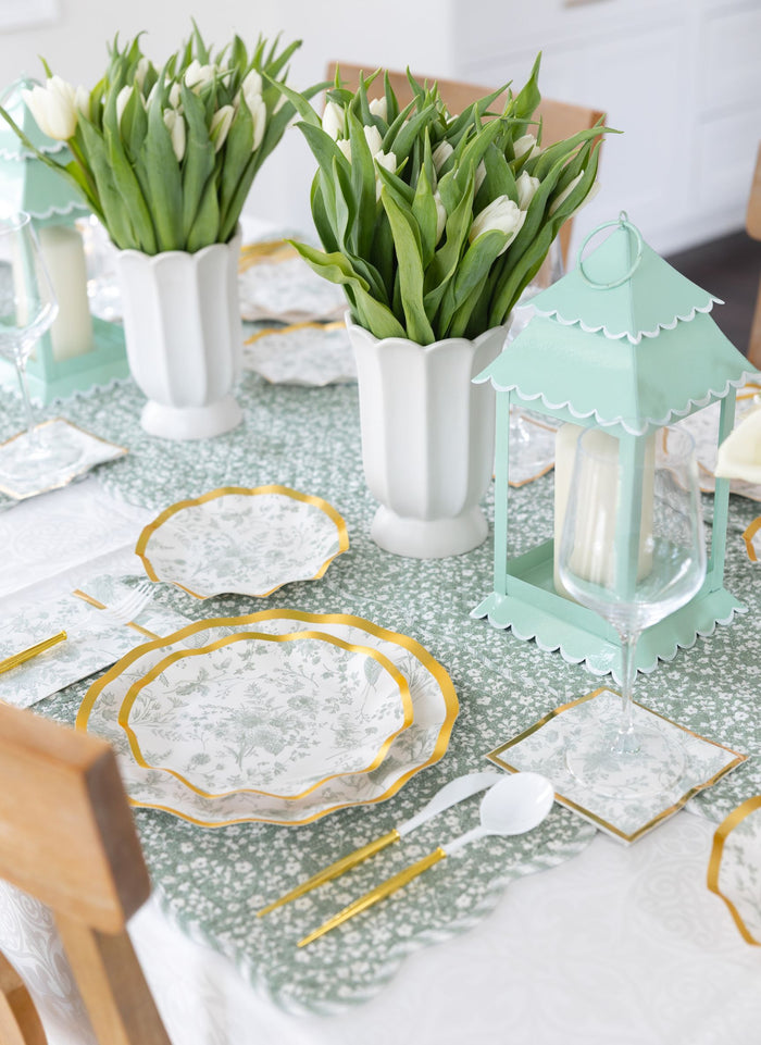 Create a spring tablescape with Sophistiplate's Timeless Table Setting: Sage—designer floral plates, gold utensils, white napkins, white tulip vases, a pastel green lantern centerpiece, and a green-and-white table runner.