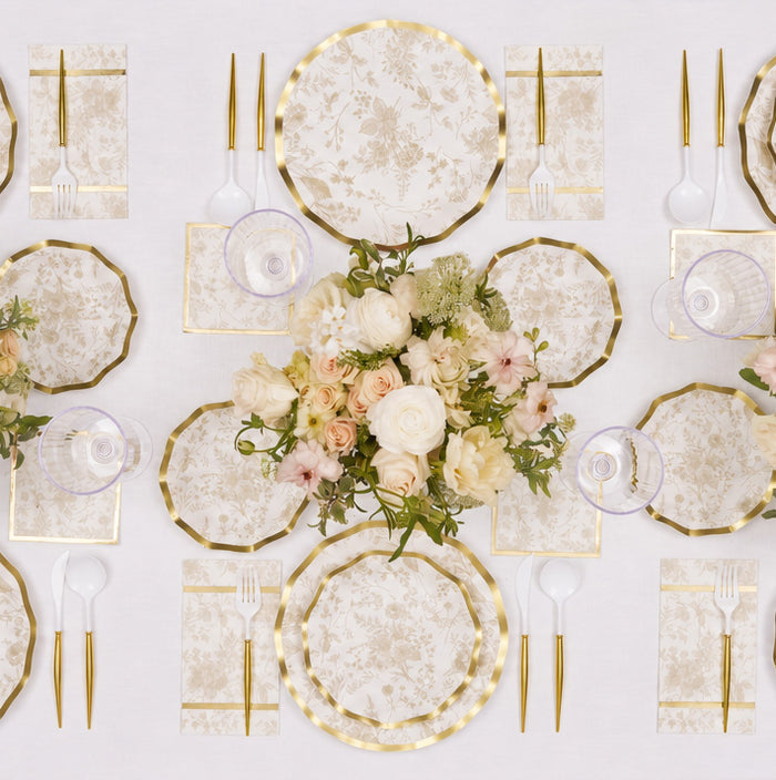Experience the Timeless Table Setting: Sand by Sophistiplate, featuring gold-rimmed designer paper plates, floral-patterned napkins, elegant gold cutlery, clear glasses, and a stunning centerpiece of white and blush flowers beautifully arranged on a crisp white tablecloth.