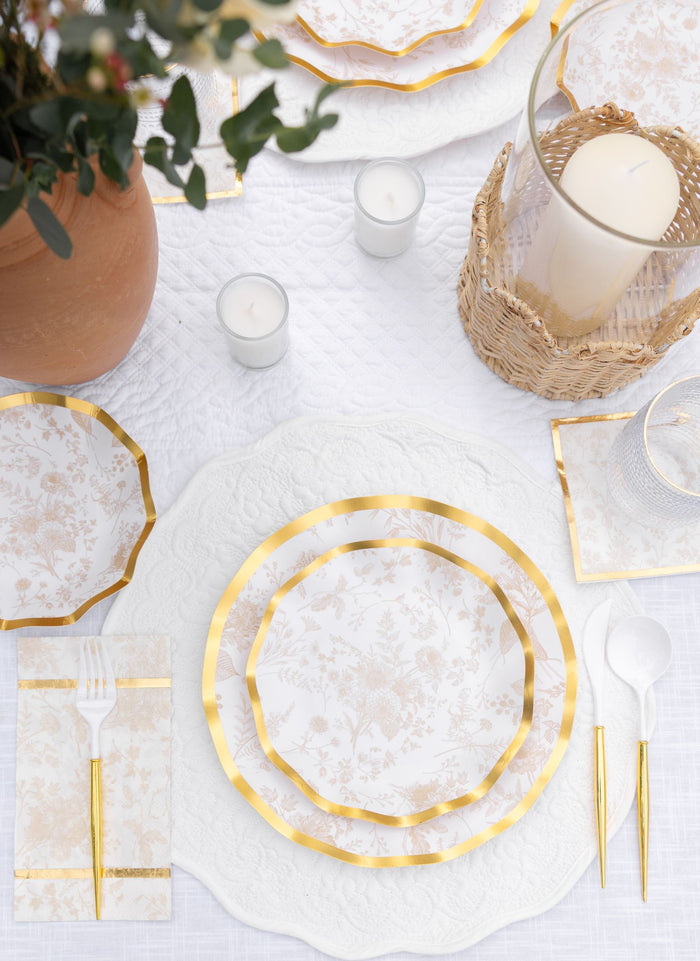 The Timeless Table Setting: Sand by Sophistiplate features a white tablecloth, gold-trimmed designer disposable plates, gold-accented cutlery, napkins, clear glassware, glass candle holders, a potted plant, and a large candle in a wicker holder.