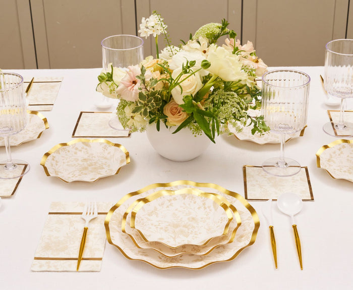 The Timeless Table Setting: Sand by Sophistiplate features designer disposable plates, gold utensils, clear glasses, and floral napkins. A white bowl with white and pale pink flowers creates an elegant centerpiece.