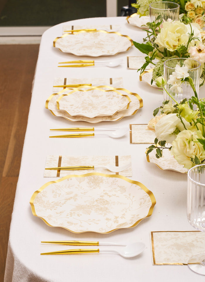 A chic table features Sophistiplate Timeless Sand Dinner Plates (8pk), gold chopsticks, white spoons, napkins, and glasses on a white tablecloth, accented with white and yellow floral arrangements.