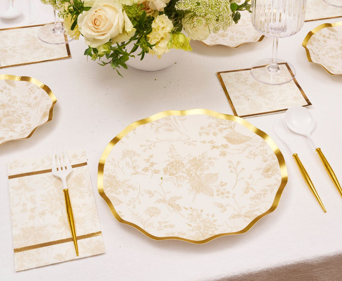 Elegant table setting with Sophistiplate Timeless Sand Dinner Plates (8pk), matching napkin, white and gold fork, glass and spoon, plus a floral centerpiece of white and cream blooms on a white tablecloth.