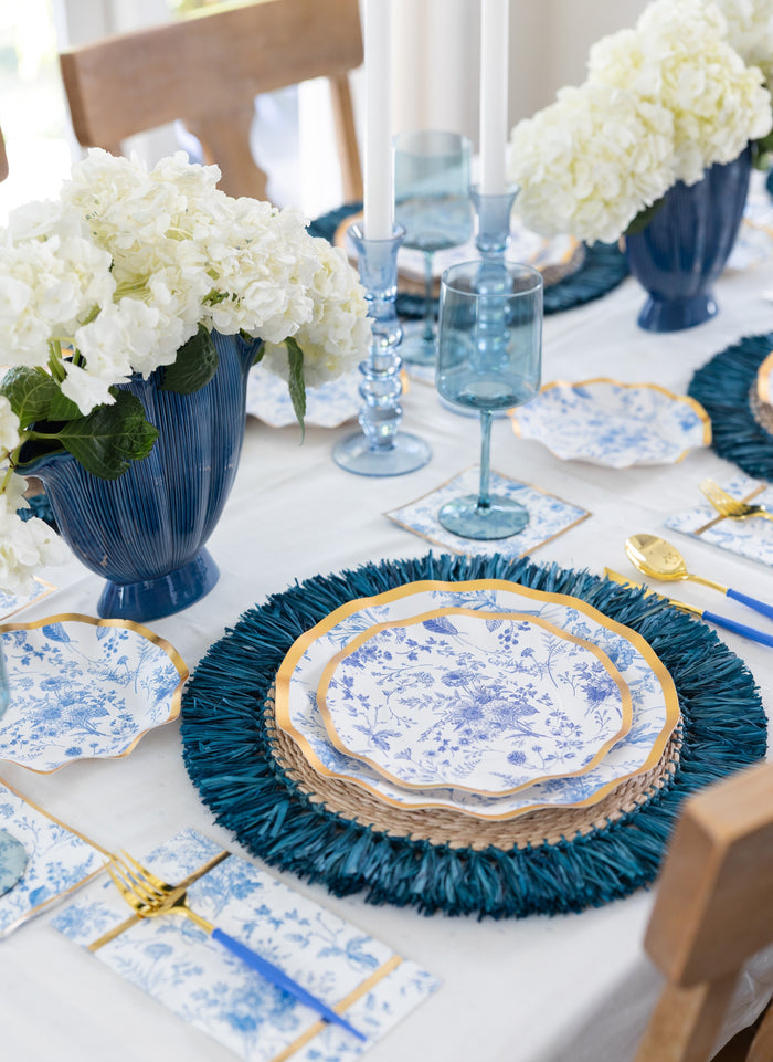 The elegant table set features blue and white floral plates, gold cutlery, blue-tinted glassware, and white hydrangea centerpieces in blue vases, beautifully enhanced by Sophistiplate’s Timeless Guest Towels (20pk) that perfectly complement the sophisticated and coordinated dining atmosphere.