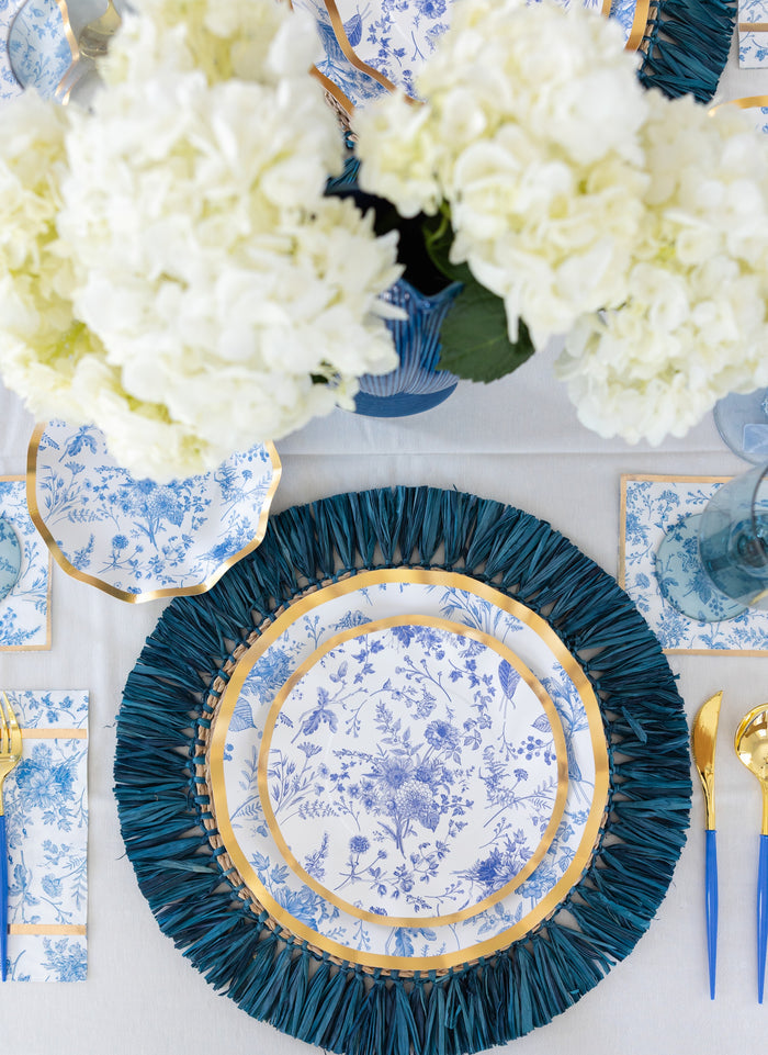 Elevate your table setting with Sophistiplate’s Timeless Guest Towels (20pk), perfectly complementing blue and white floral plates with gold-rimmed edges, gold flatware, blue flowery napkins, and white flower centerpieces atop a crisp white tablecloth. A blue placemat adds a vibrant touch of color.