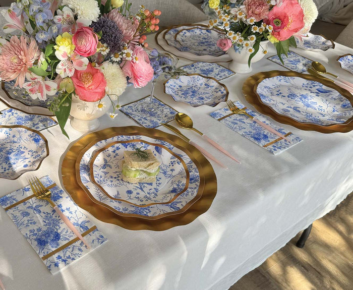 A sophisticated table setting showcases Sophistiplate’s Timeless Dinner Plates (8pk), featuring a blue and white floral design with a metallic gold rim. Gold and pink cutlery, elegant floral centerpieces, and petite appetizers set atop each plate enhance the look, all arranged on a crisp white tablecloth. Sunlight gently illuminates the scene, creating an inviting ambiance ideal for any holiday celebration.