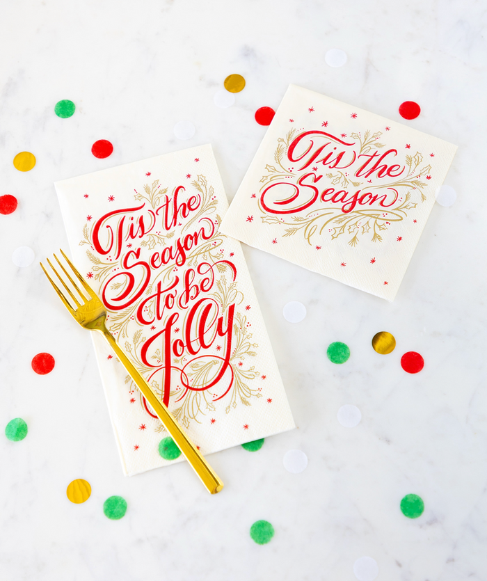 Hester & Cook’s ‘Tis the Season Cocktail Napkins (20pk) are displayed with a gold fork and confetti on a white marble surface, along with a festive card featuring “’Tis the Season to be Jolly” in red and gold script.