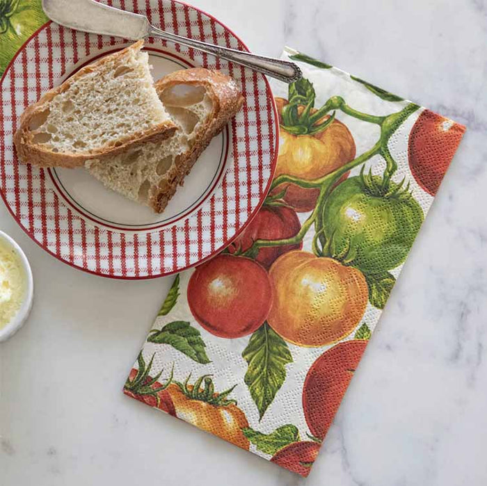 A plate with two slices of bread and a butter knife sits next to Hester & Cook Tomato Guest Napkin (pack of 16) on a marble surface, with a small dish of butter partially in view.
