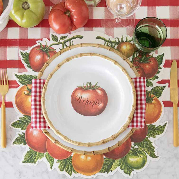 A tomato-themed table setting features Hester & Cook’s Tomato Place Card - Pack of 12, a white plate with a tomato illustration and the name Mario, red checkered napkin, gold cutlery, green glasses, and fresh tomatoes on a red checkered tablecloth.