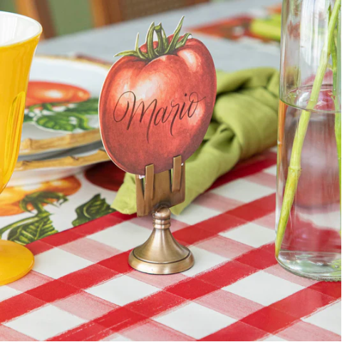 The Hester & Cook Tomato Place Card - Pack of 12 sits on a metal holder atop a checkered tablecloth, with a yellow glass, green napkin, and vase—ideal as eco-friendly buffet labels for your next gathering.